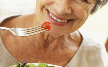 Starsza kobieta zkrótkimi siwymi włosami z napitym na widelec pomidorkiem koktailowym, uniesionym nad talerzem z sałatką ze świeżych warzyw.